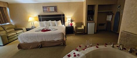 A hotel room with a large bed, a sitting area with a chair, a desk, a minibar, a bathroom with a bathtub, and a shower.