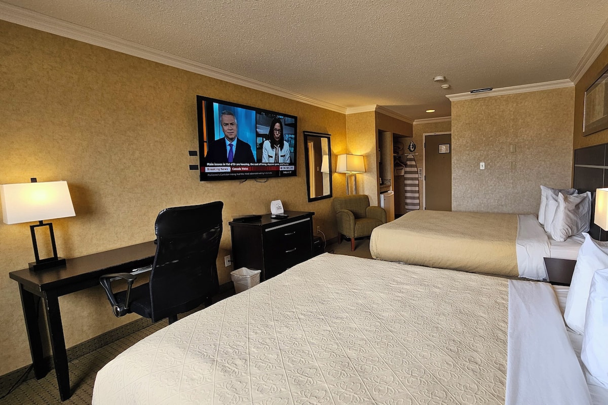 A hotel room with two beds, a desk, a chair, a television displaying news, and a lamp.