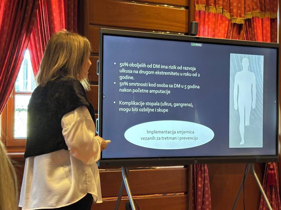 A person presenting information on a screen in a room with wooden paneling and red curtains.