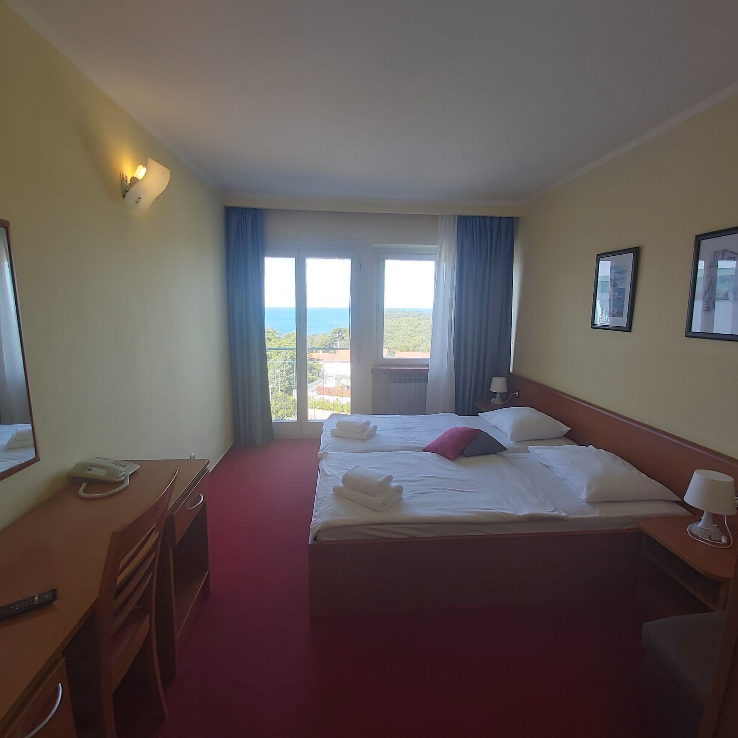 A hotel room with a bed, a desk, a phone, and a view of the sea.