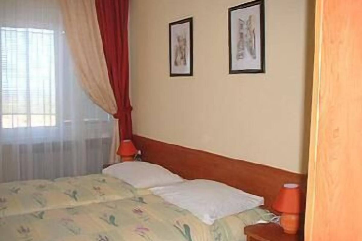 Una cama doble con ropa de cama floral, un cabecero de madera, dos cuadros enmarcados en la pared y una ventana con cortinas traslúcidas.