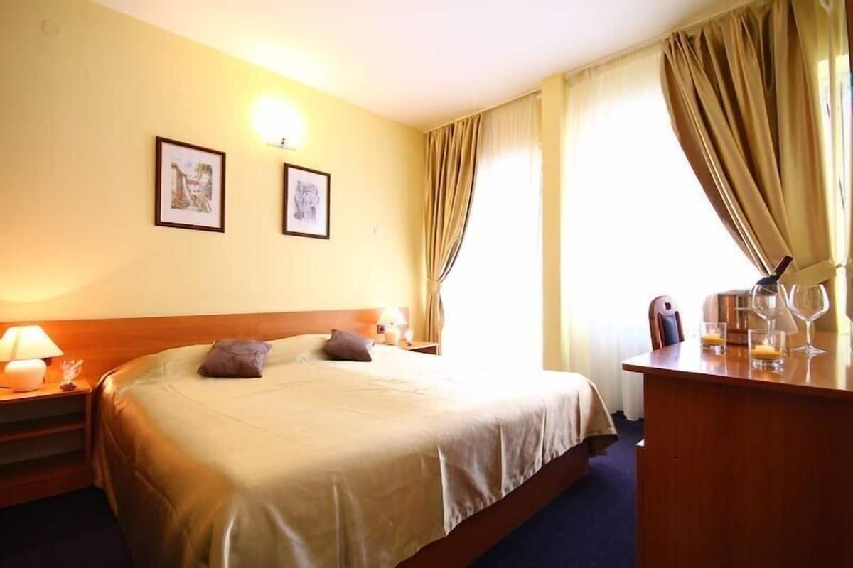 Una habitación de hotel con cama, mesitas de noche, un escritorio con vasos y una lámpara, y cortinas.