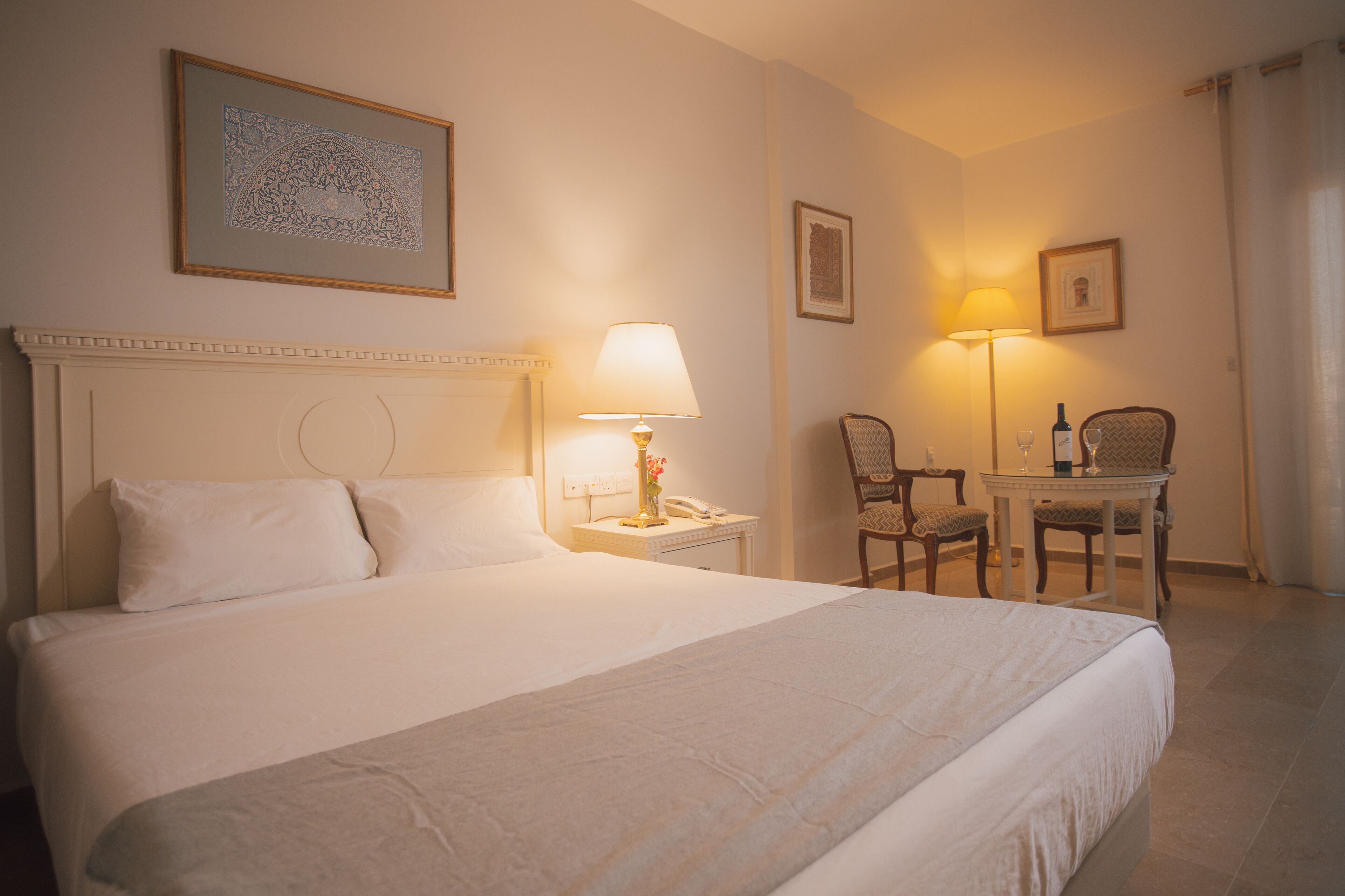 A hotel room with a large bed, a bedside table with a lamp, a chair, and a small table with a bottle and glass.