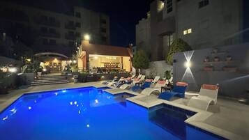 Zona de piscina nocturna con sillones de descanso y un bar.