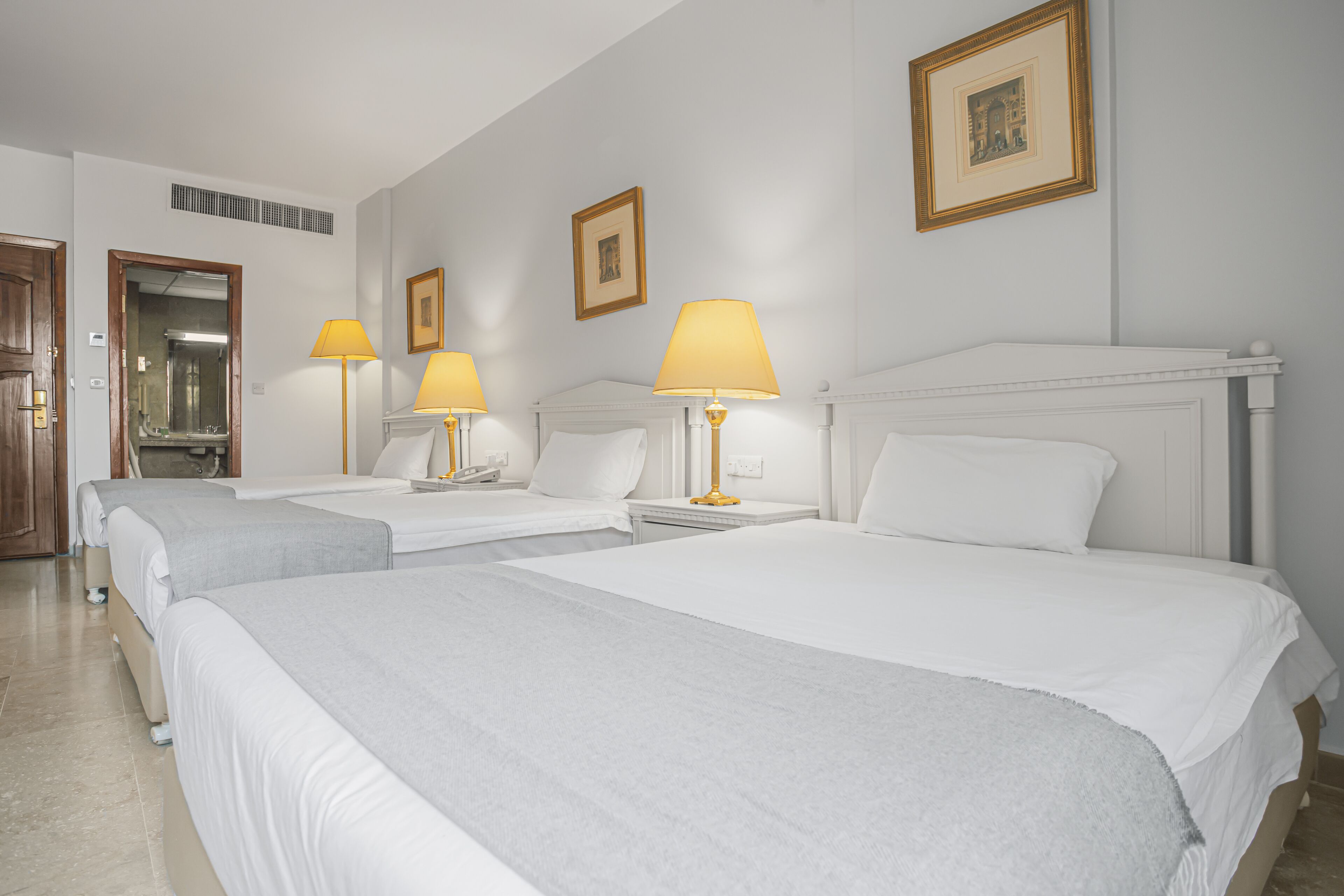 Een hotelkamer met drie bedden, elk voorzien van een witte spreidek en een lamp. Er hangen afbeeldingen in lijsten aan de muur en een deur met een spiegel.