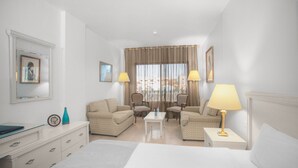 A hotel room with a bed, two armchairs, a small table, and a nightstand with a lamp. There is a window with curtains and a framed picture on the wall. - Grand Hotel Madaba (Madaba)