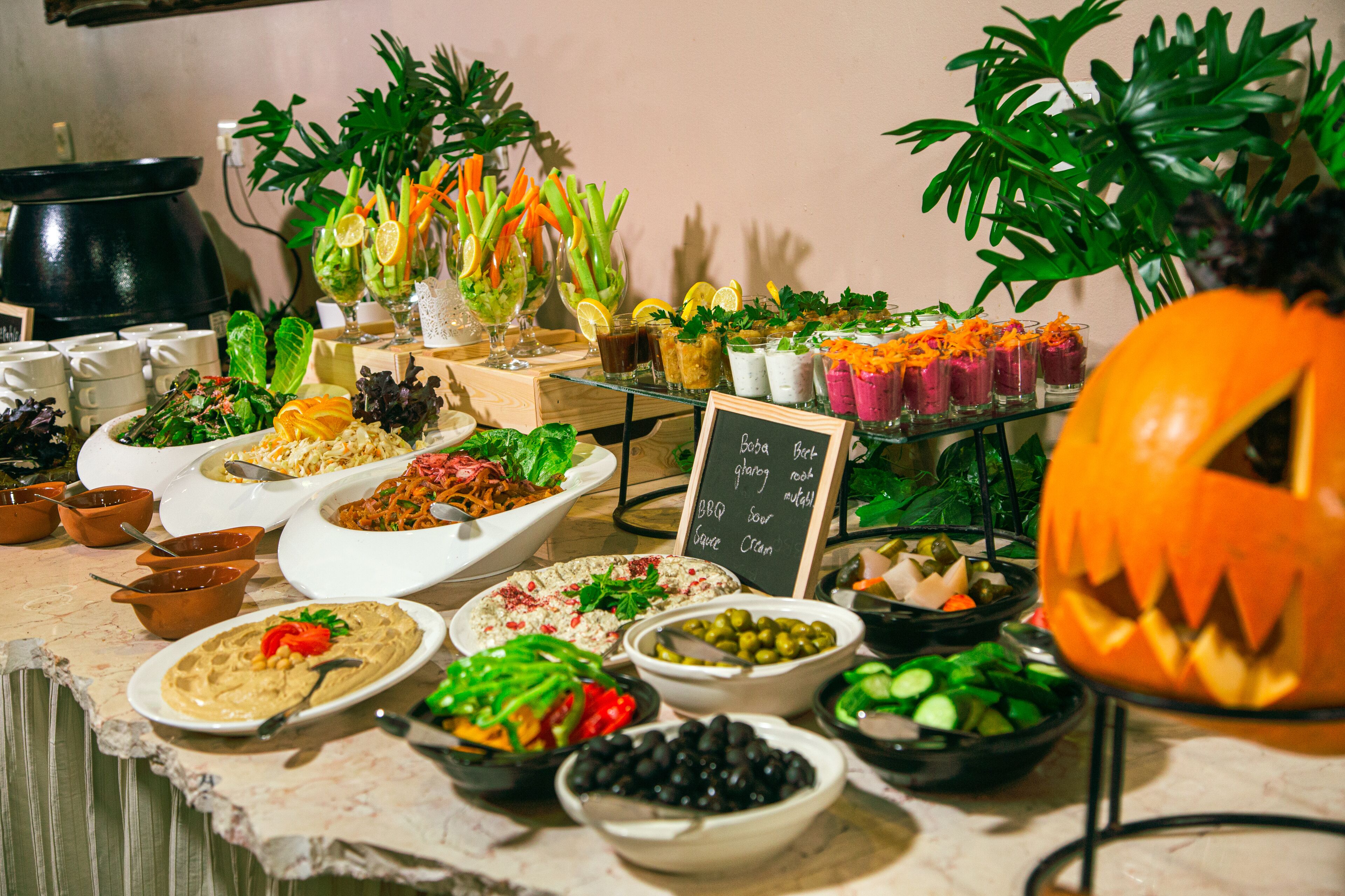 Een buffettafel met diverse gerechten en drankjes, waaronder salades, dips en een uitgesneden pompoen.