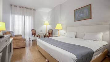 Ein Hotelzimmer mit einem großen Bett, zwei Nachttischen inklusive Lampen, einem Sessel, einem Schreibtisch und einem dekorativen Wandbild.