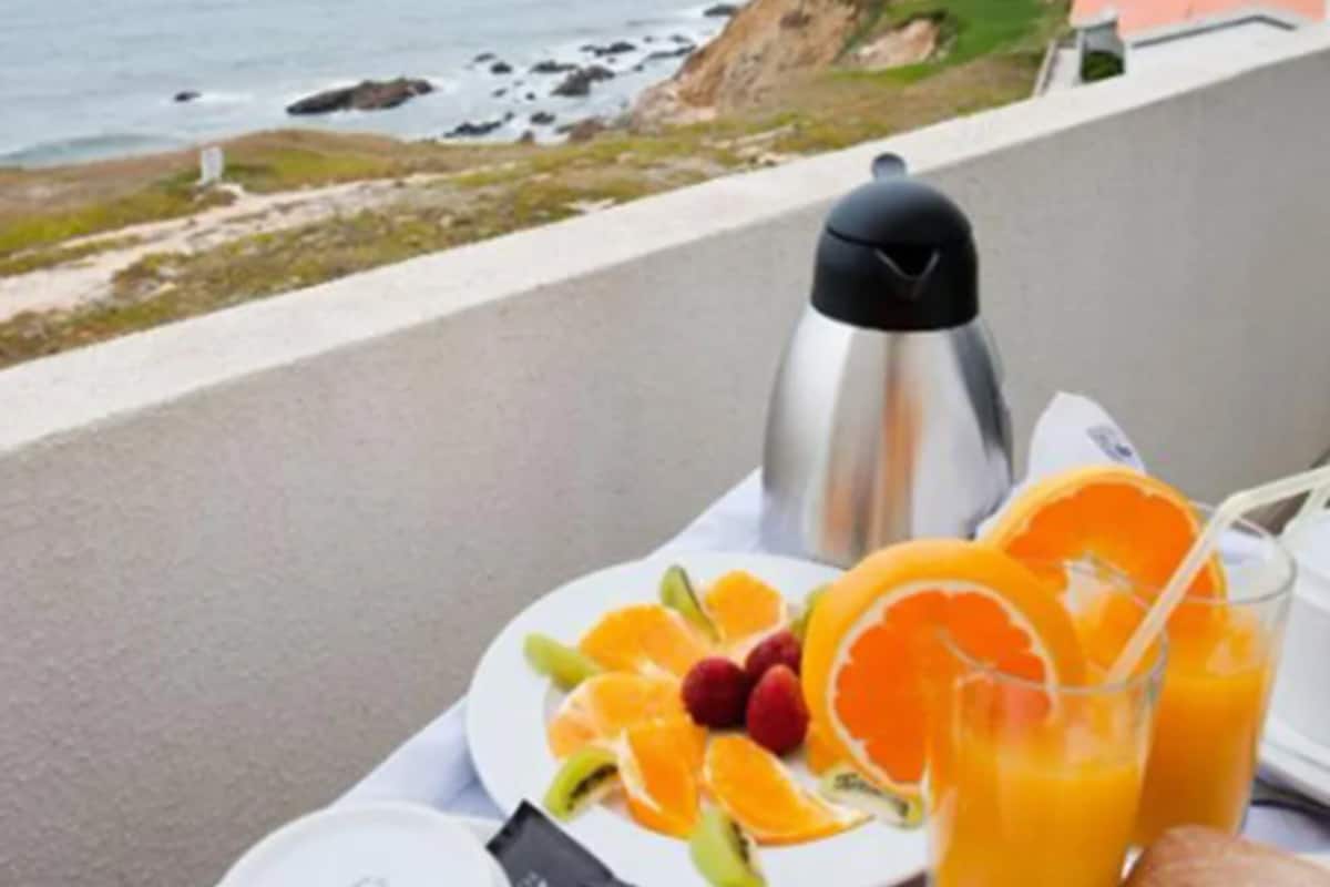 A breakfast setup on a balcony overlooking the sea, featuring a plate of fruit, juice glasses, a coffee pot, and a croissant.