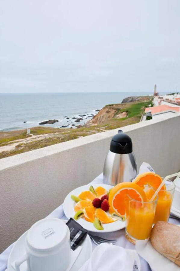 A breakfast setup on a balcony overlooking the sea, featuring a plate of fruit, juice glasses, a coffee pot, and a croissant.
