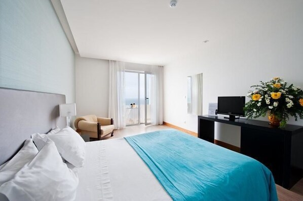 A hotel room with a bed, a chair, a desk with a TV, and a view of the outdoors. - Hotel Mar e Sol & Spa (Marinha Grande)