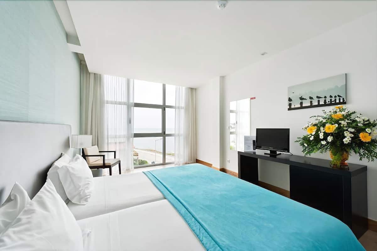 A hotel room with a bed, a desk with a TV, a chair, a window with curtains, and a painting on the wall.