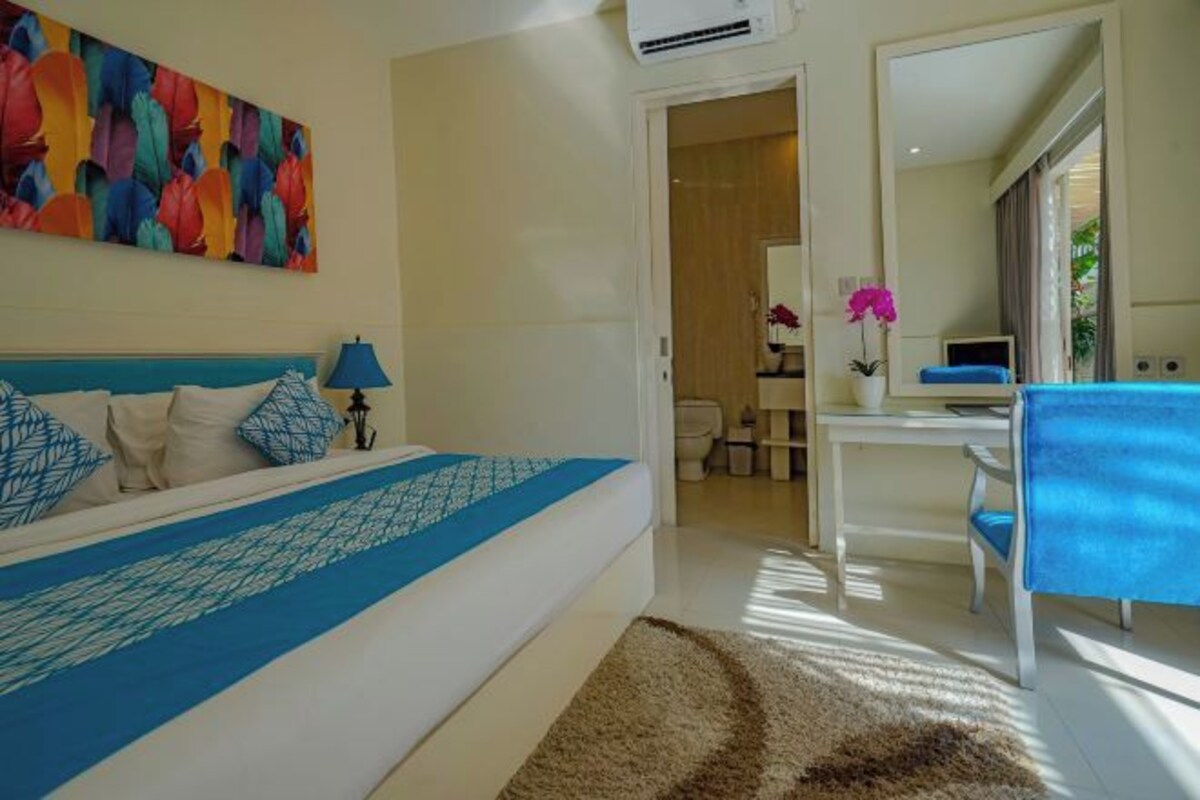Une chambre d’hôtel avec un lit, un bureau, une chaise et une salle de bain visible à travers une porte ouverte.