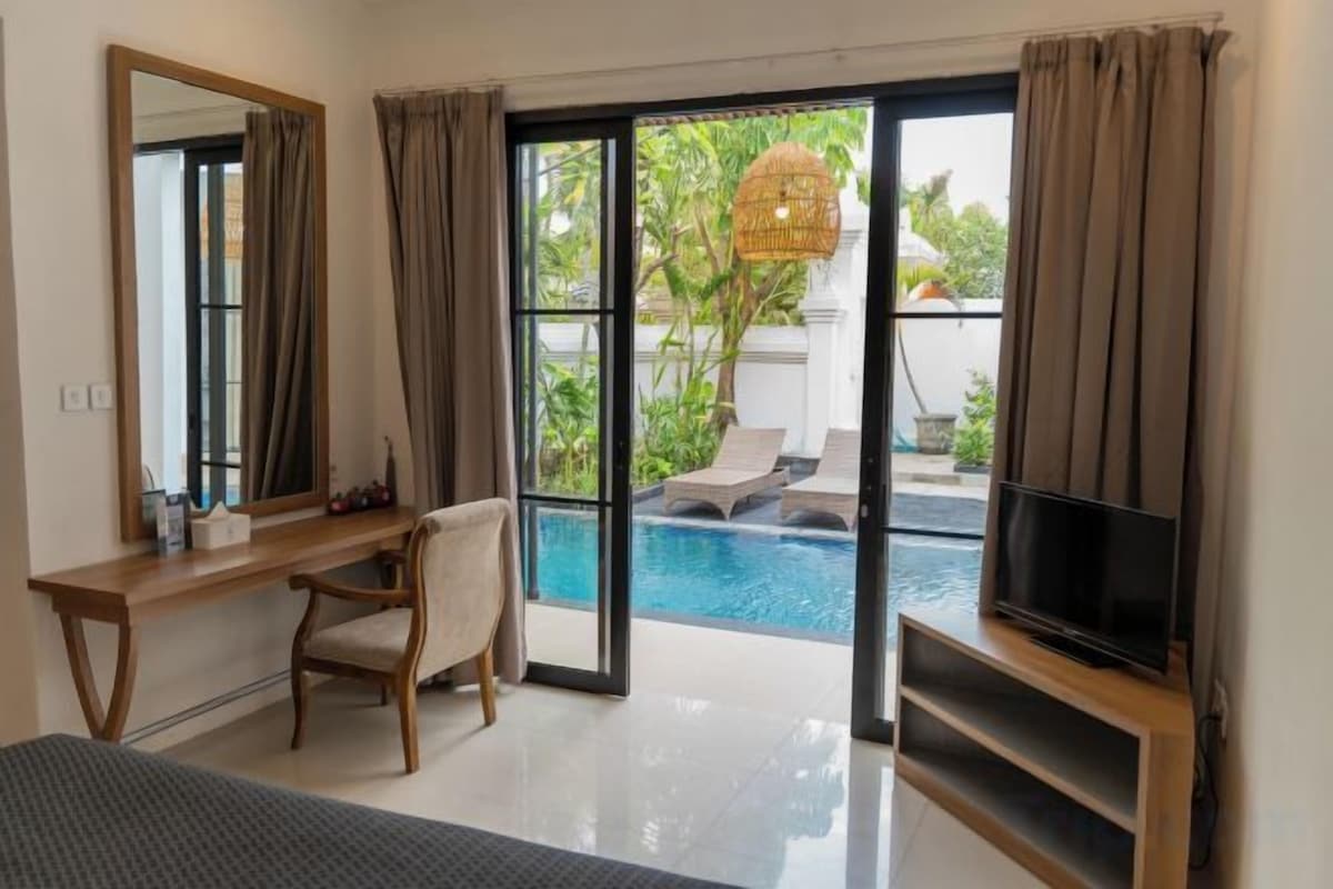 Un dormitorio con cama, escritorio, silla, televisión y vista a una piscina al aire libre.