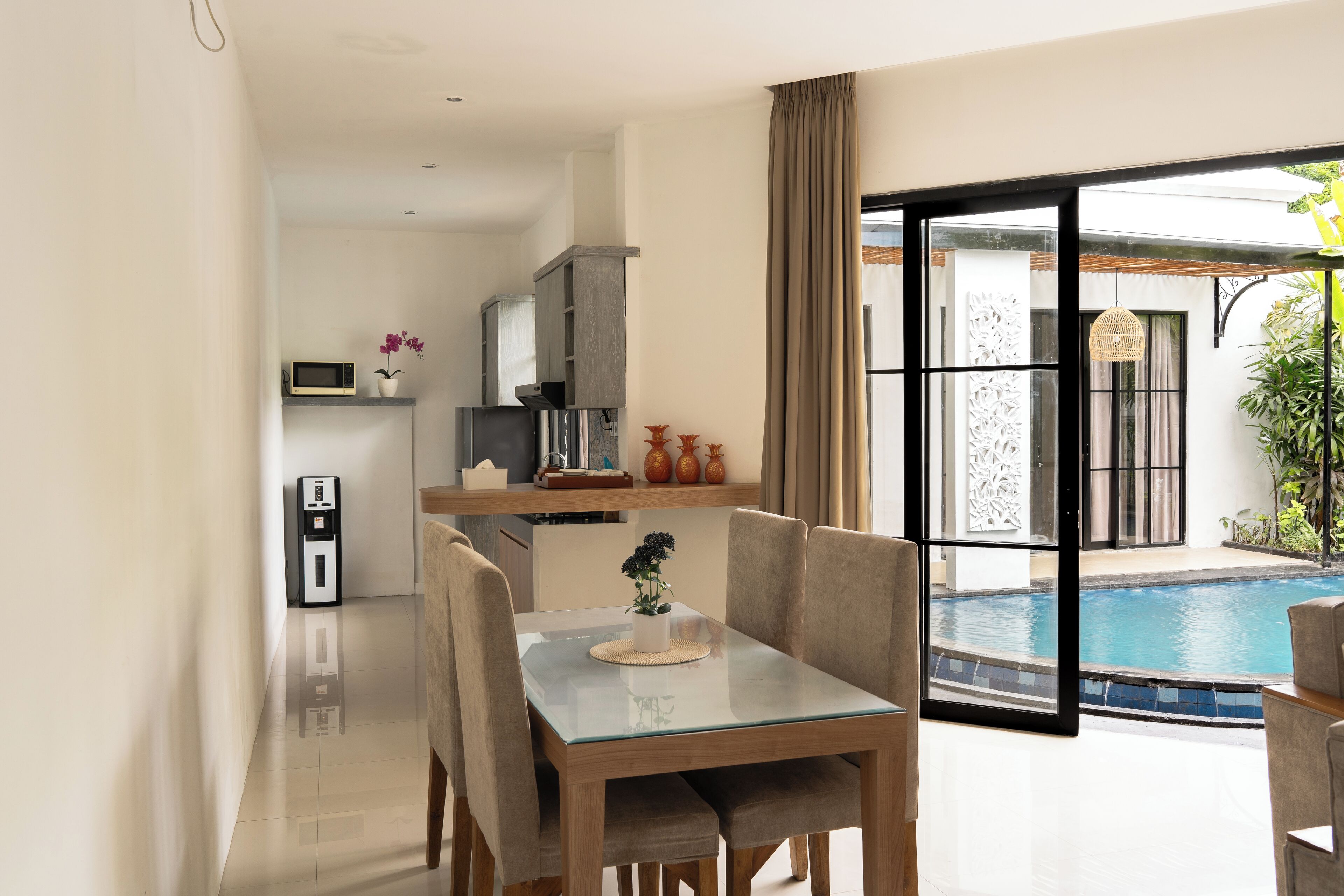 Un comedor moderno con una mesa y sillas de vidrio, una cocina al fondo y vista a una piscina.