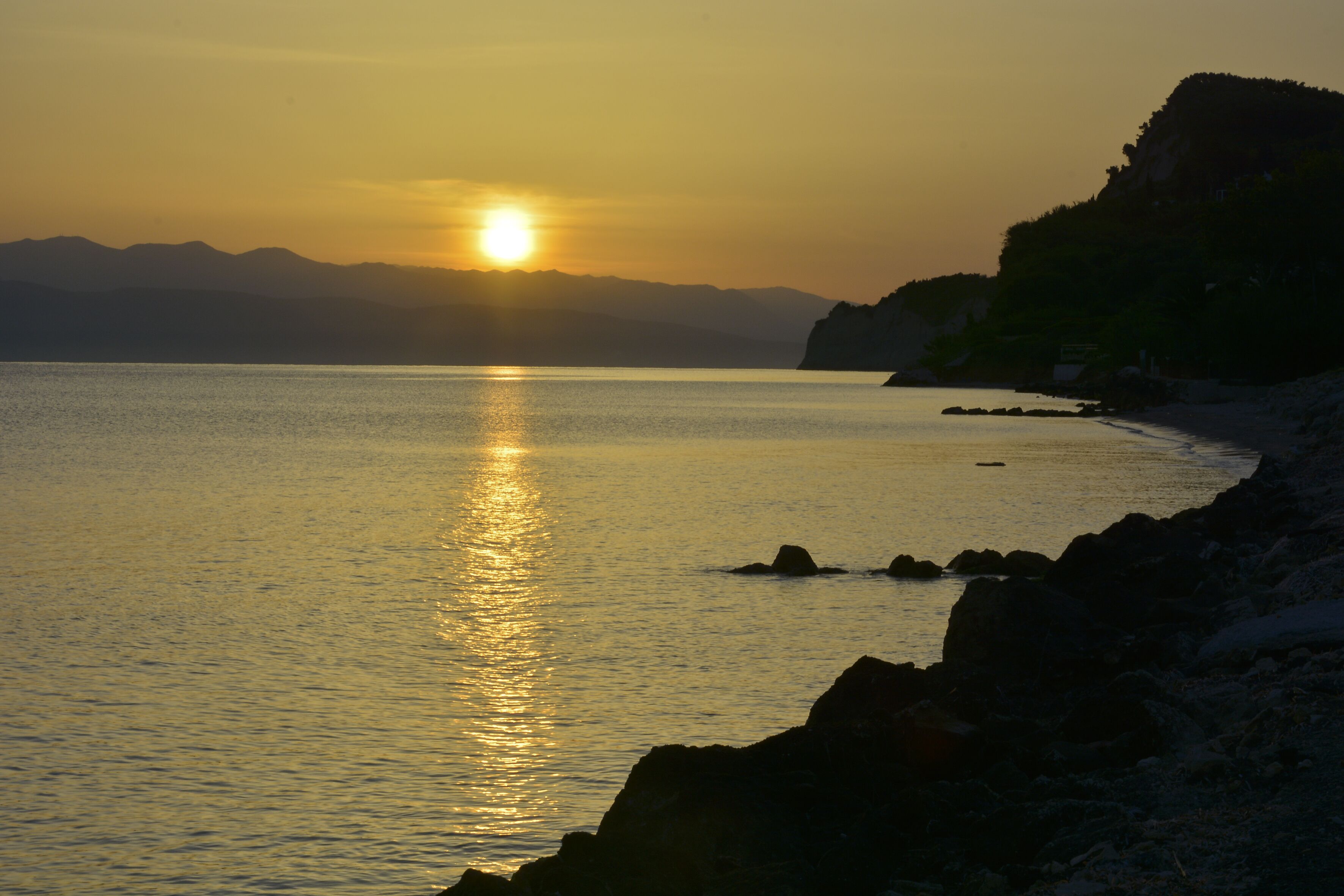 A sunset over a calm sea with rocky shores and distant mountains.