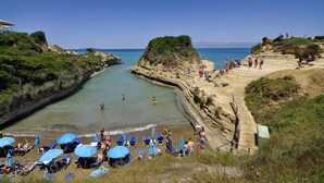 A beach with clear waters, rocky cliffs, and people enjoying the sun. - Sidari Beach Hotel (Corfu)