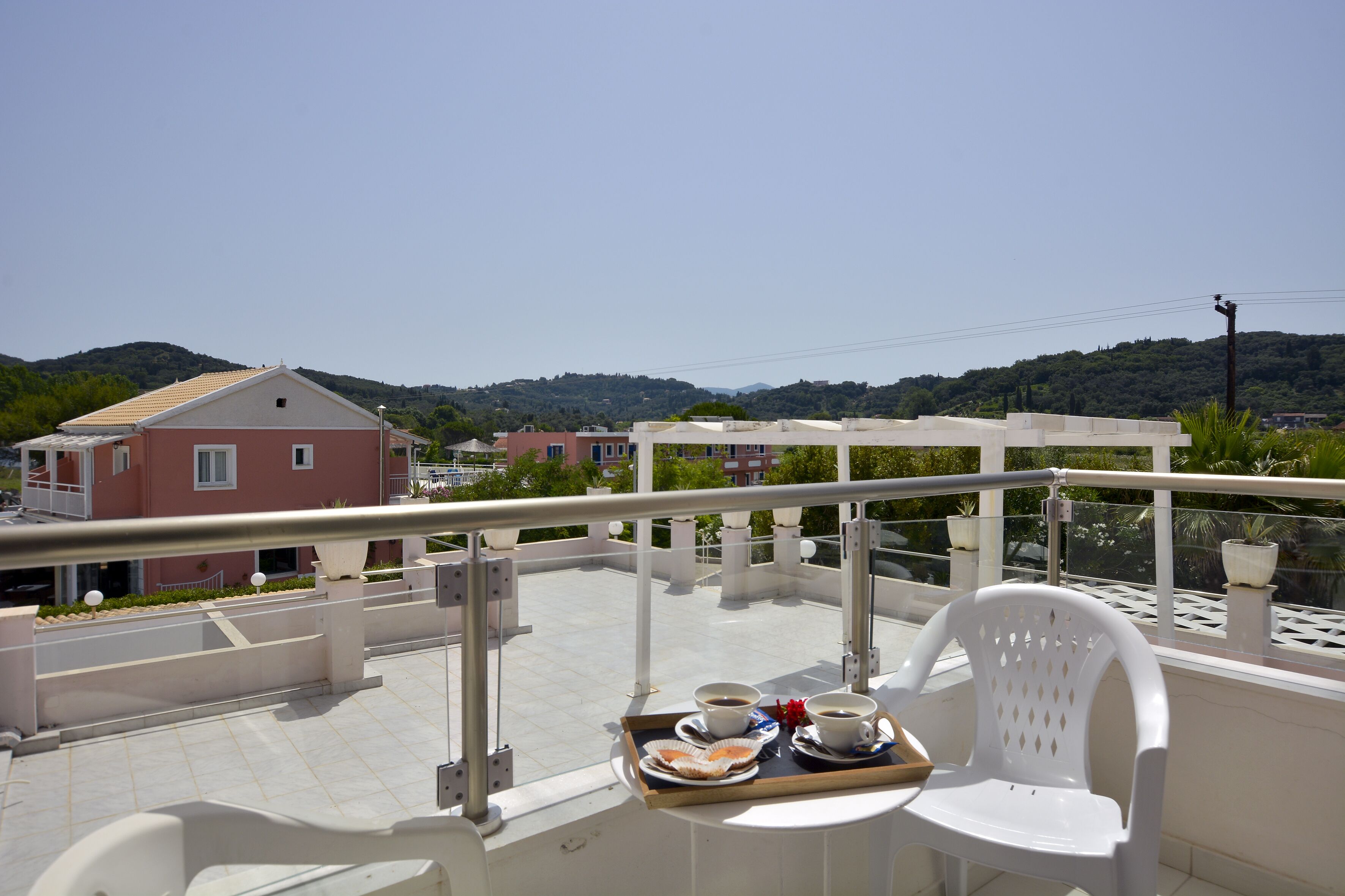 A balcony with a white plastic chair, a tray with a cup of coffee and a pastry, and a view of the surrounding buildings and hills.