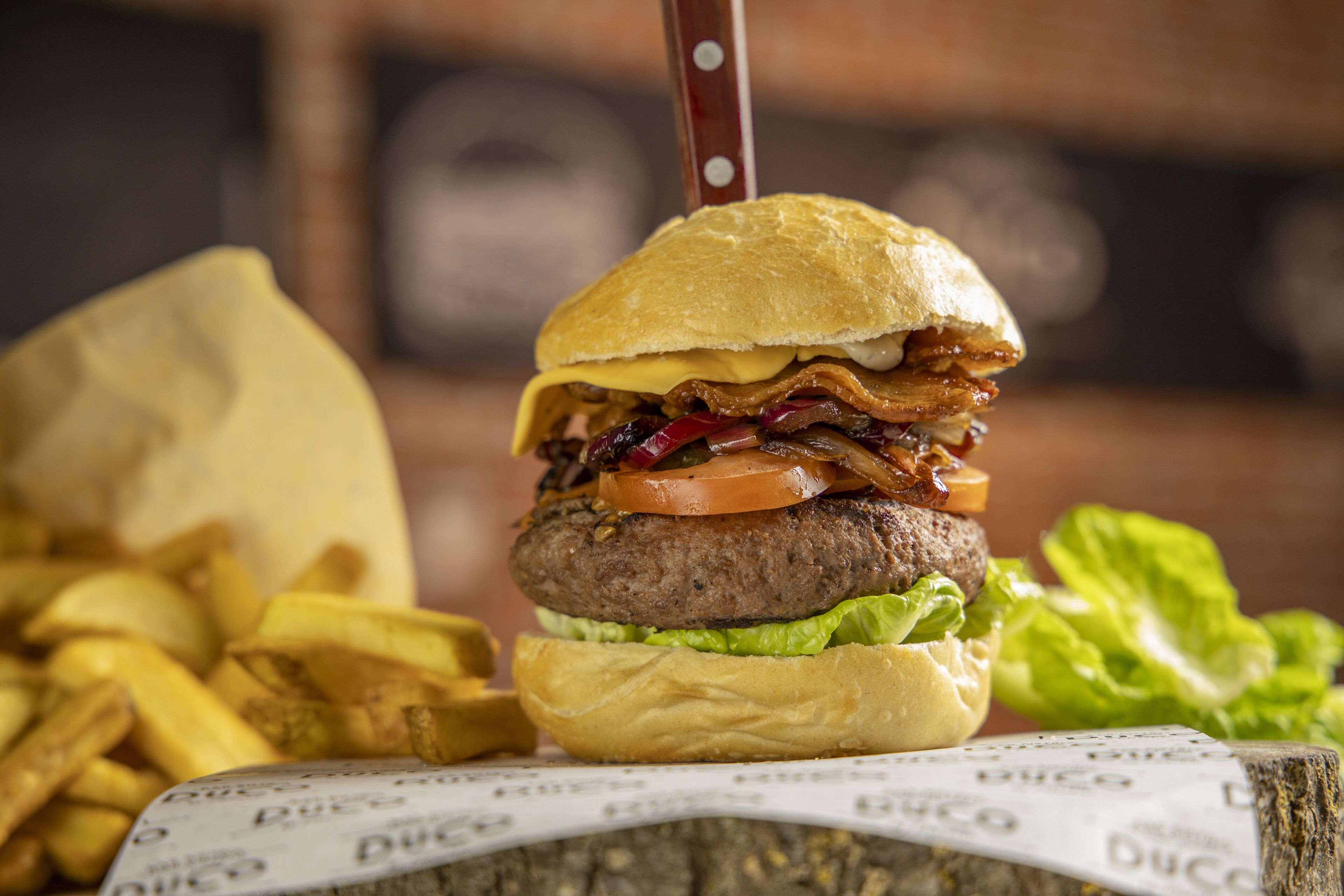 Um hambúrguer com bacon, tomate e alface no pão, acompanhado de batatas fritas e uma faca.