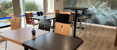 A modern dining area with wooden tables and chairs, a large window with blue tape, and a mural of tropical foliage on the wall.