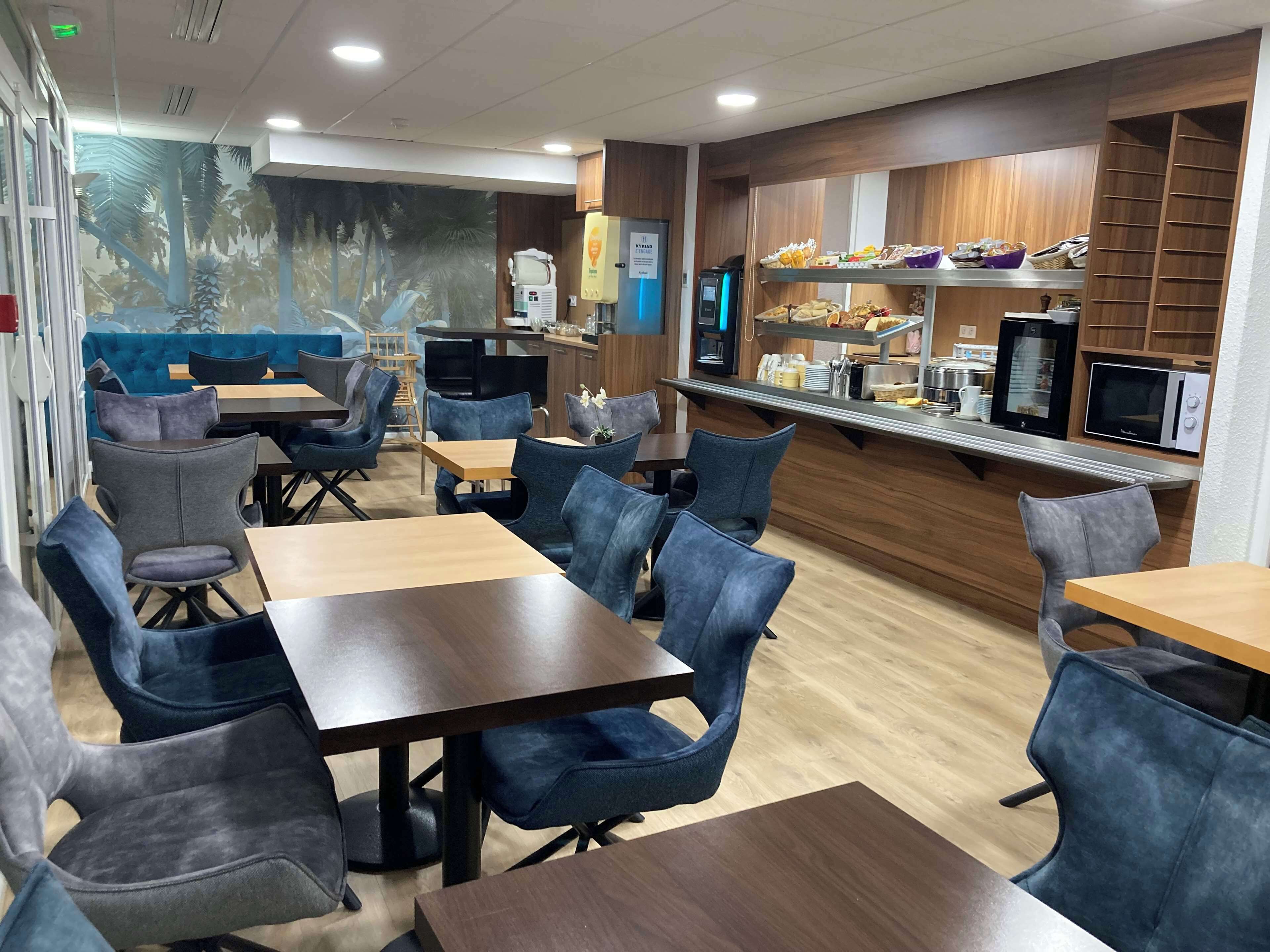 A restaurant with wooden tables and blue chairs, a kitchen area with appliances, and a mural on the wall.