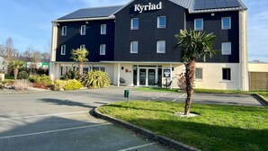 A modern hotel named Kyriad with a dark facade, multiple windows, and a parking area in front.