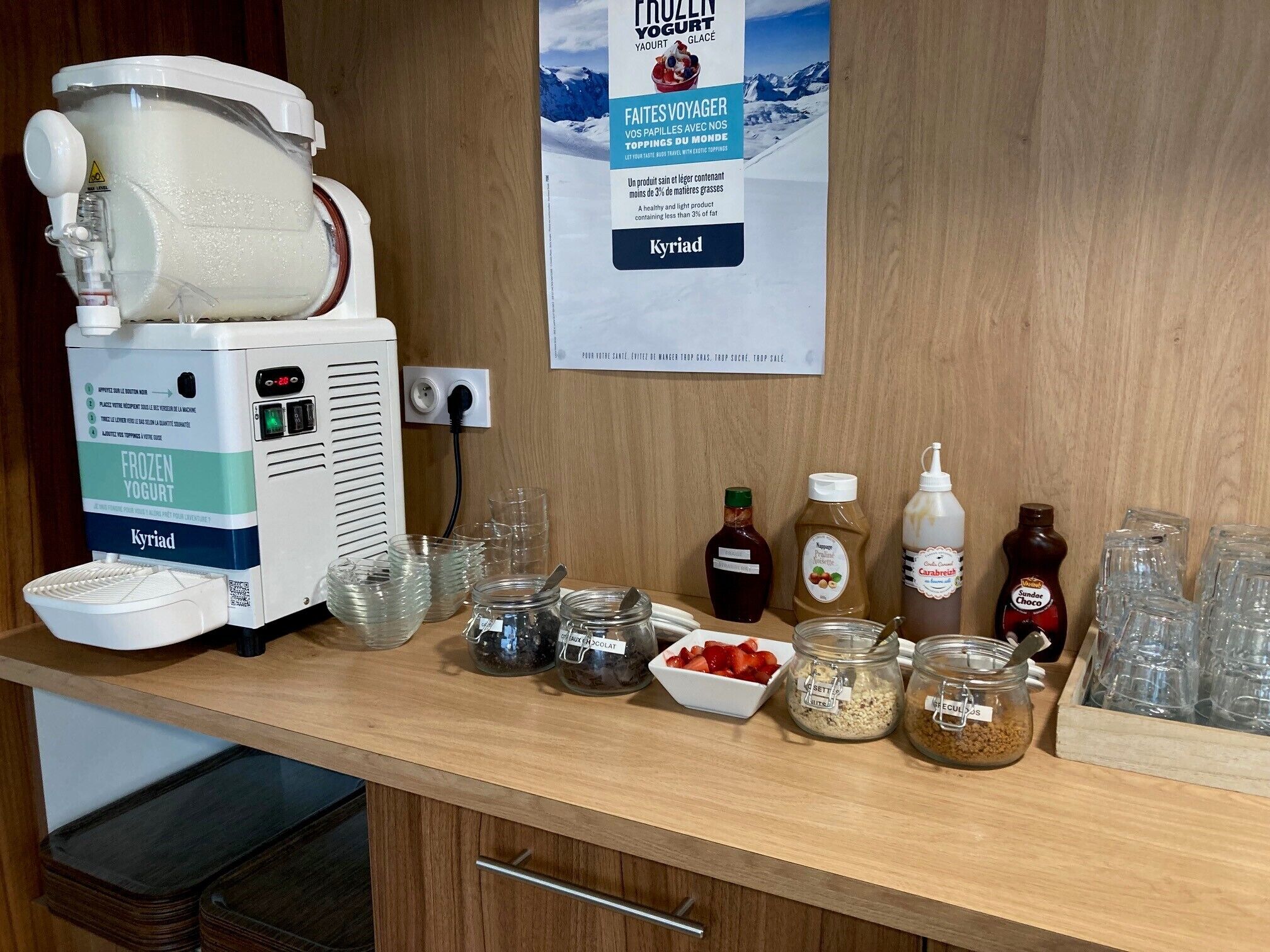 A frozen yogurt machine, various condiments, and glass containers on a wooden counter.
