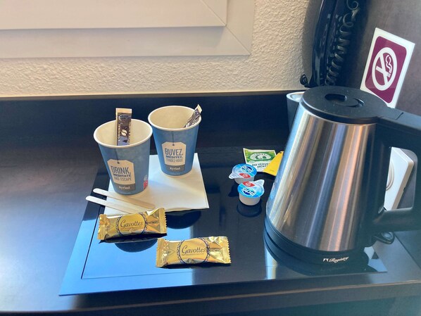 Una estación de café con dos tazas etiquetadas 'DRINK, MEDITATE AND ESCAPE', una cafetera de acero inoxidable y dos paquetes de chocolate con leche Gavottes.