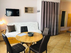 A cozy room with a sofa, a TV mounted on the wall, a small wooden shelf, and a dining table with chairs.