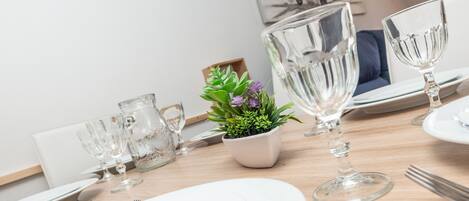 Una mesa de comedor dispuesta con platos blancos, cristalería transparente y una pequeña maceta con flores moradas.