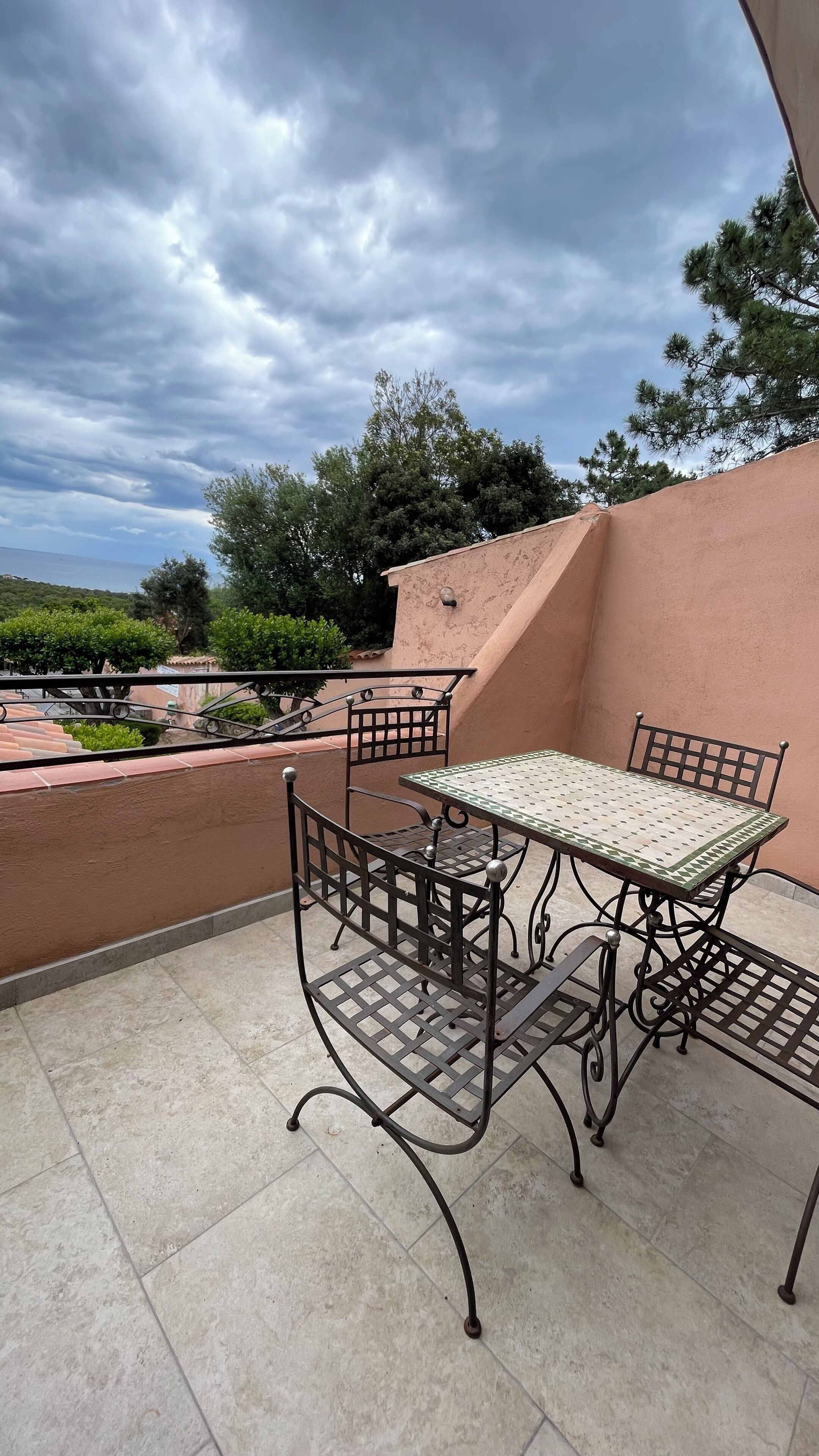 A patio with a metal table and chairs, a tiled floor, and a view of the sea and trees under a cloudy sky.