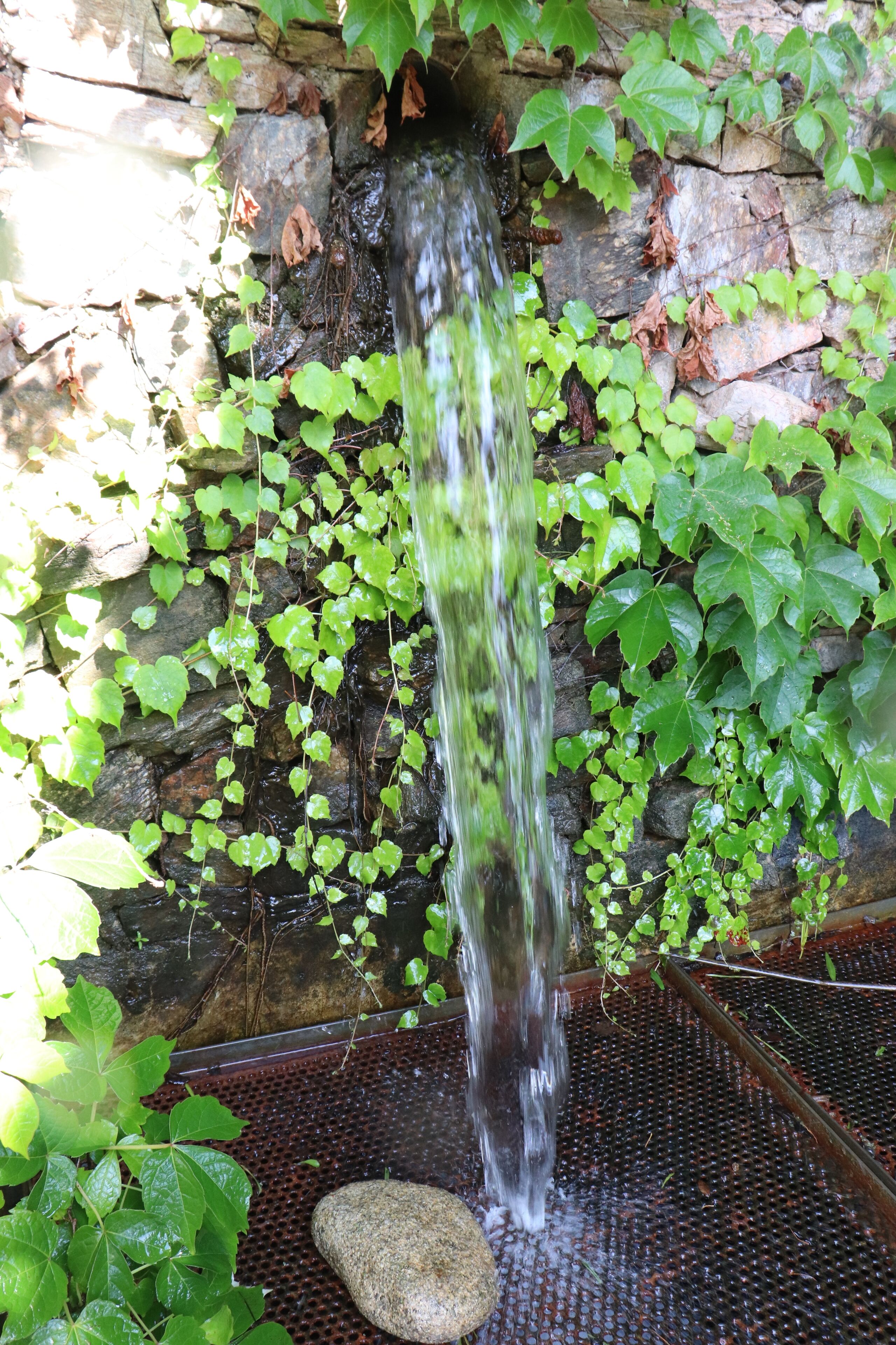 A waterfall cascading over a stone wall, surrounded by green foliage.