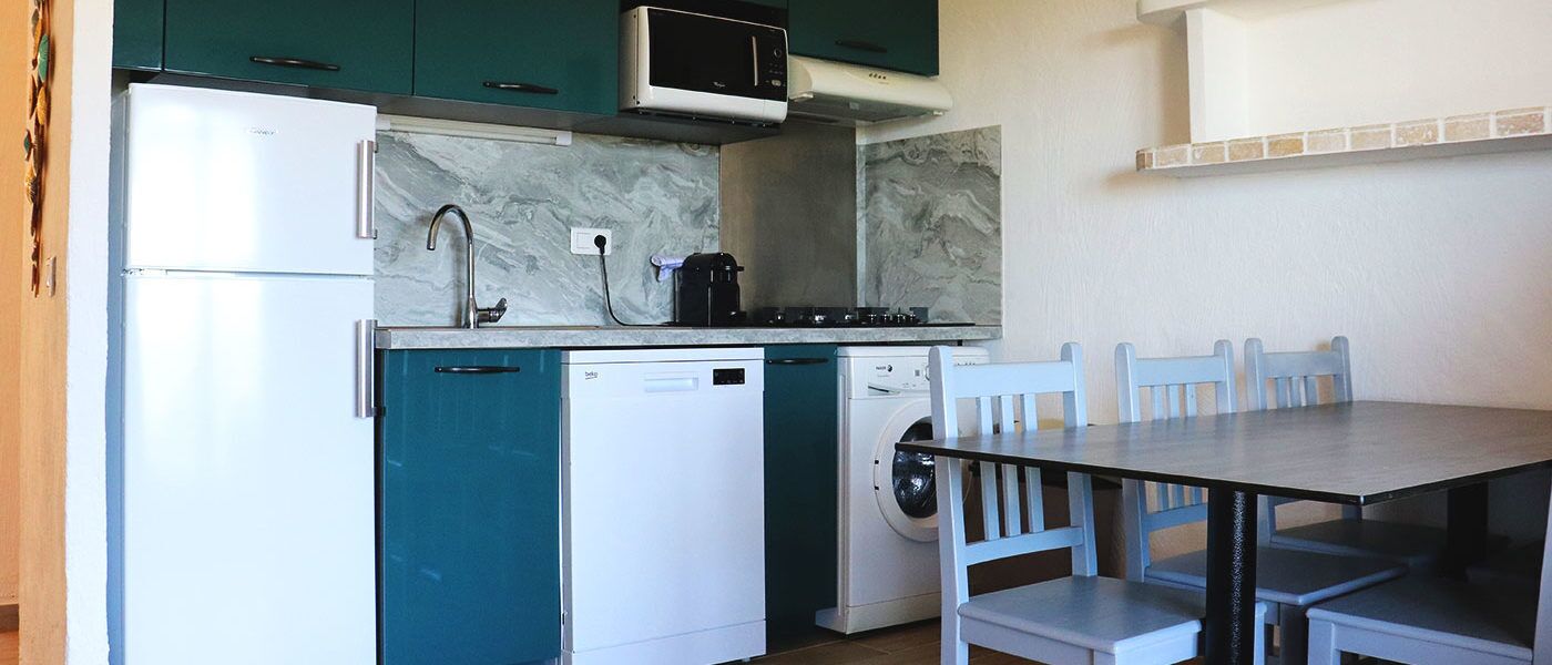 A compact kitchen with a refrigerator, dishwasher, washing machine, and a microwave. There is a dining table with chairs.