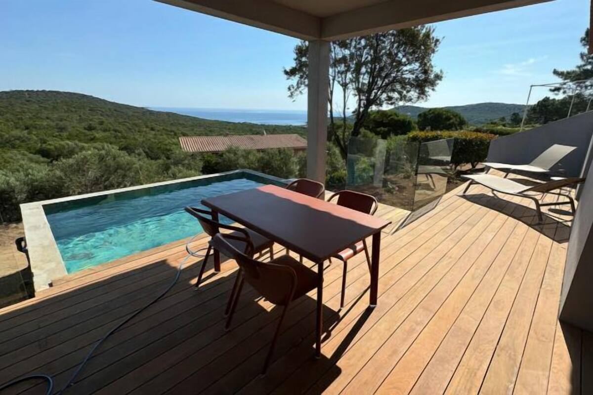 Eine Holzterrasse mit Pool, Gartenmöbeln und Blick auf das Meer und die Hügel.