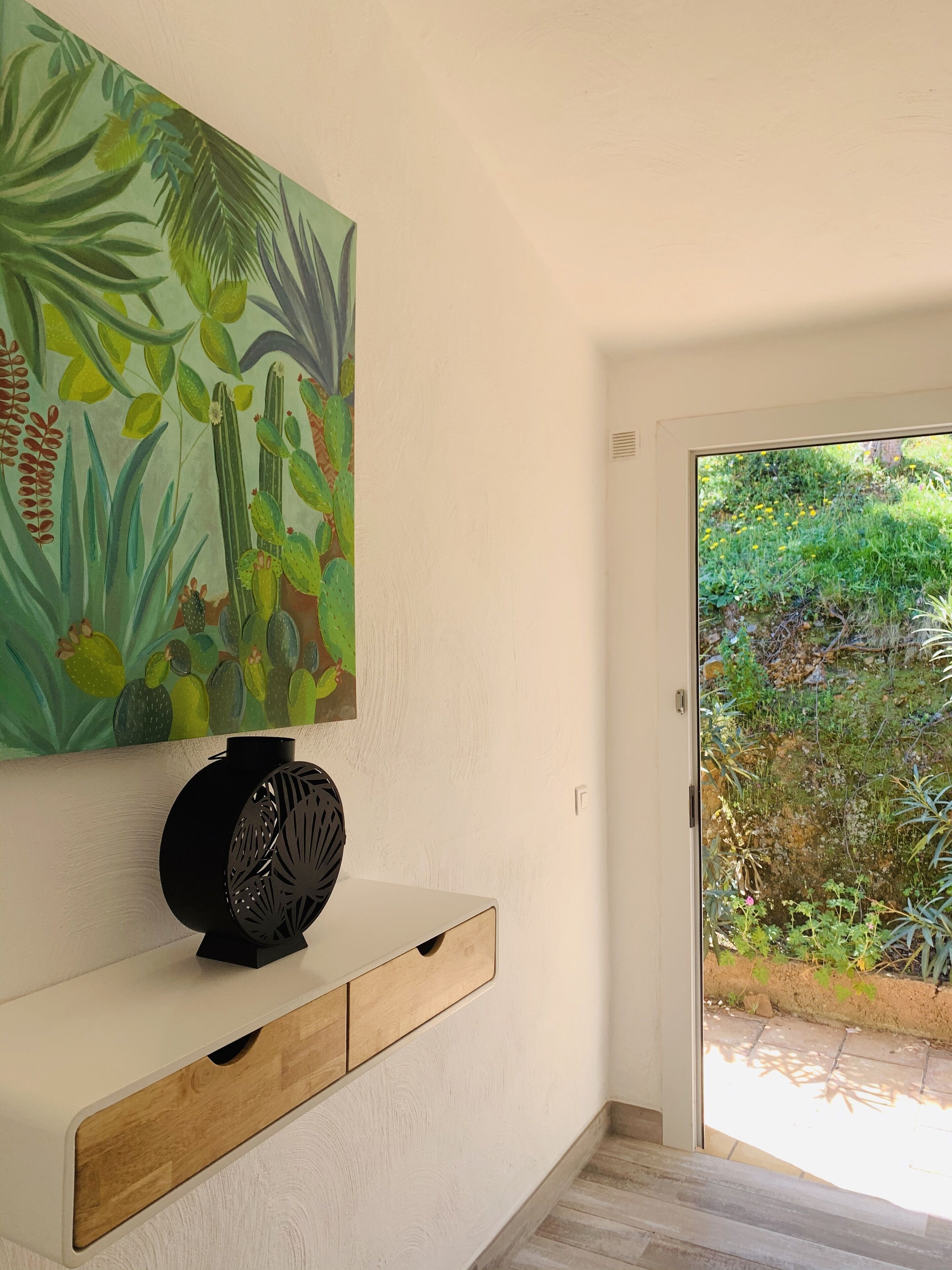 A room with a white wall, a wooden console table, a black decorative object, and a painting of tropical plants.