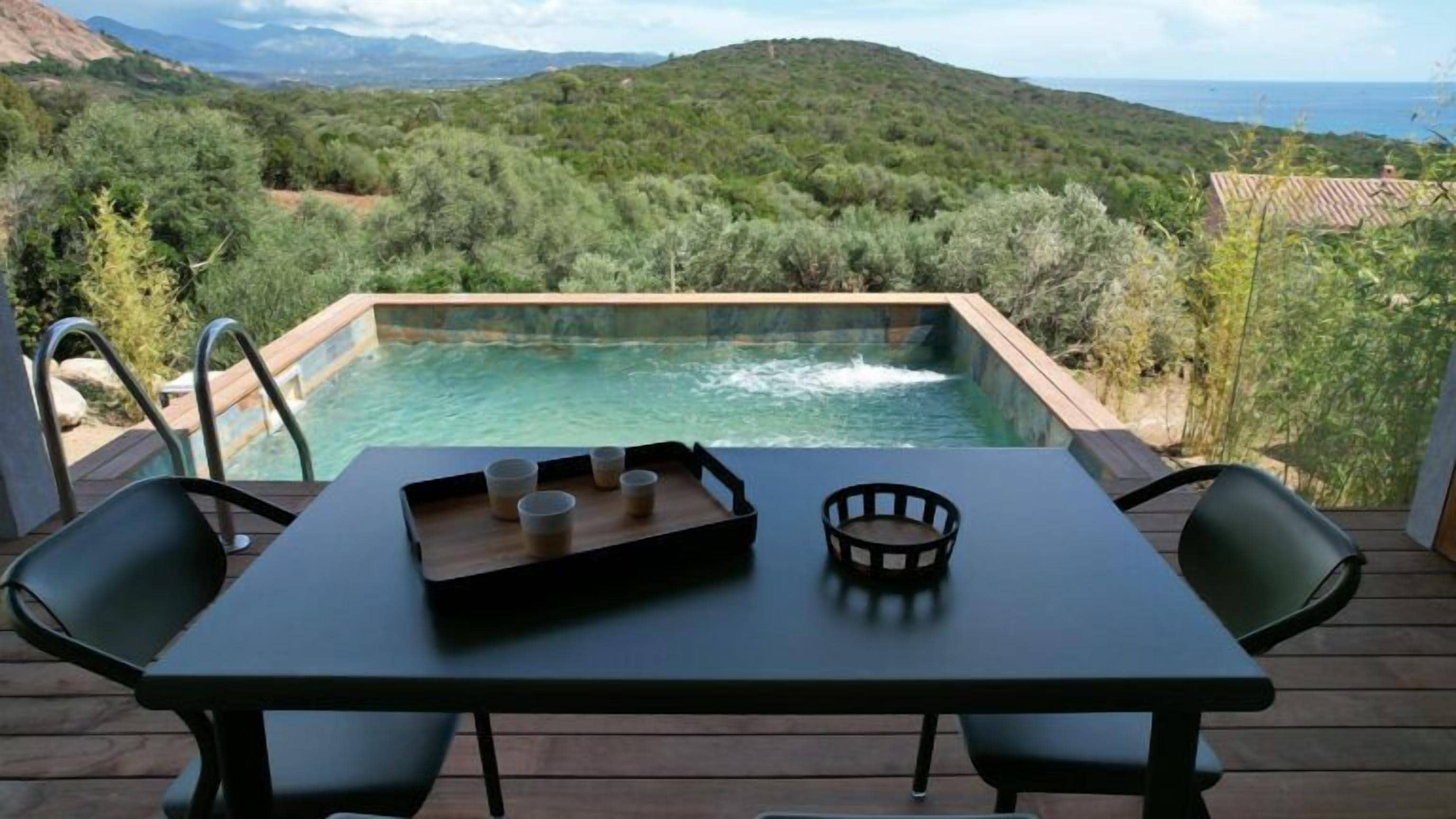 Una piscina con cubierta de madera, dos sillas, una mesa con bandeja y tazas, y vista a un paisaje montañoso.