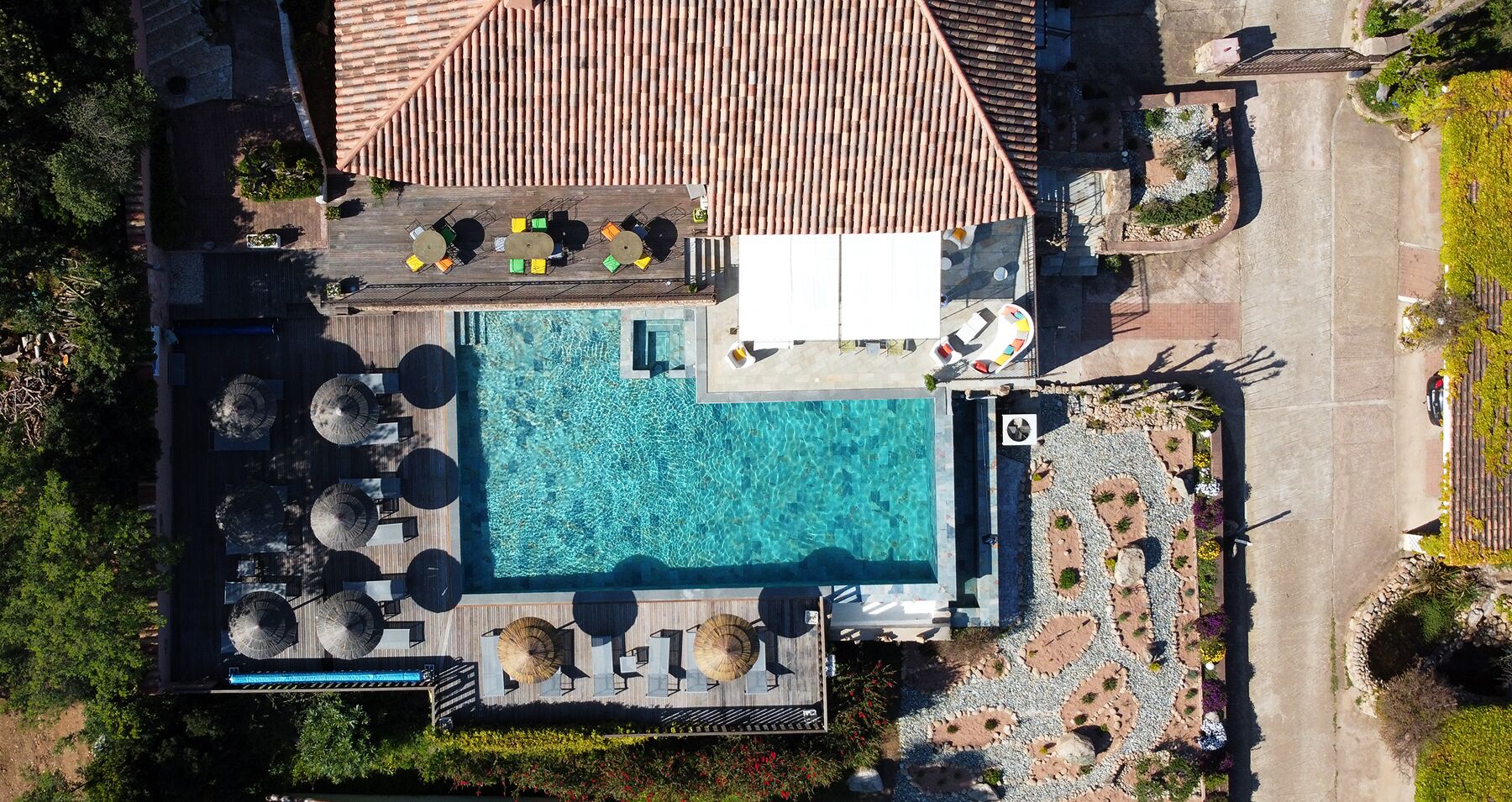 An aerial view of a hotel with a rectangular swimming pool surrounded by lounge chairs and umbrellas, a covered seating area, and a landscaped garden with pathways.