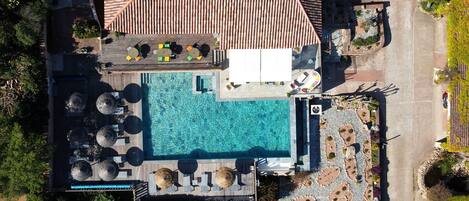 An aerial view of a hotel with a rectangular swimming pool surrounded by lounge chairs and umbrellas, a covered seating area, and a landscaped garden with pathways.