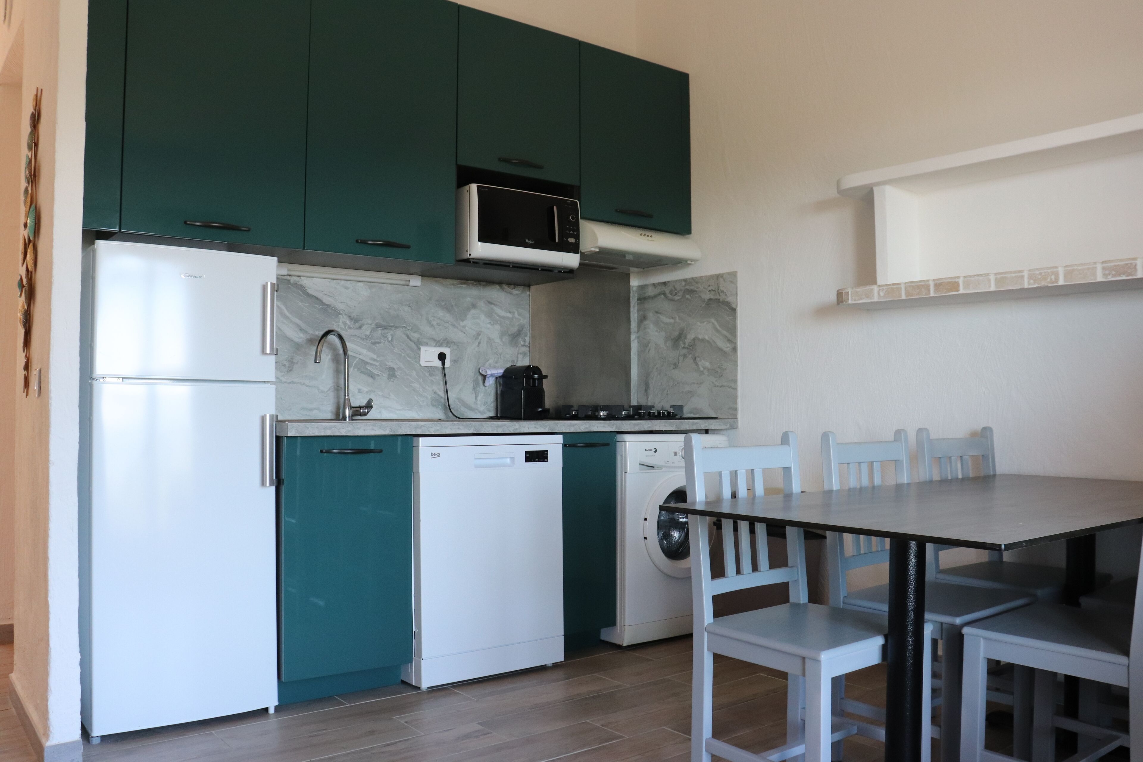 Cuina compacta amb armaris verds maragda, nevera blanca, microones, rentaplats i rentadora. Hi ha una taula de dinar amb caires i una paret blanca amb una estant a la part superior.