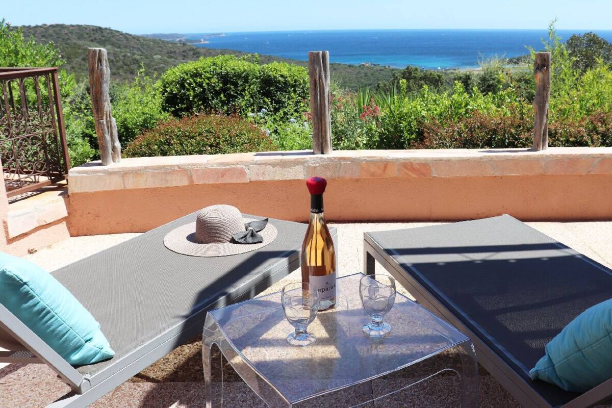 Eine Terrasse mit zwei Liegestühlen, einer Weinflasche und zwei Weingläsern auf einem Tisch, mit Blick auf eine schöne Landschaft aus Meer und Hügeln.