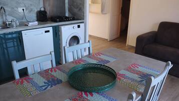 A compact kitchen with a washing machine, a dining table with a patterned table runner, and a brown sofa in the living area.