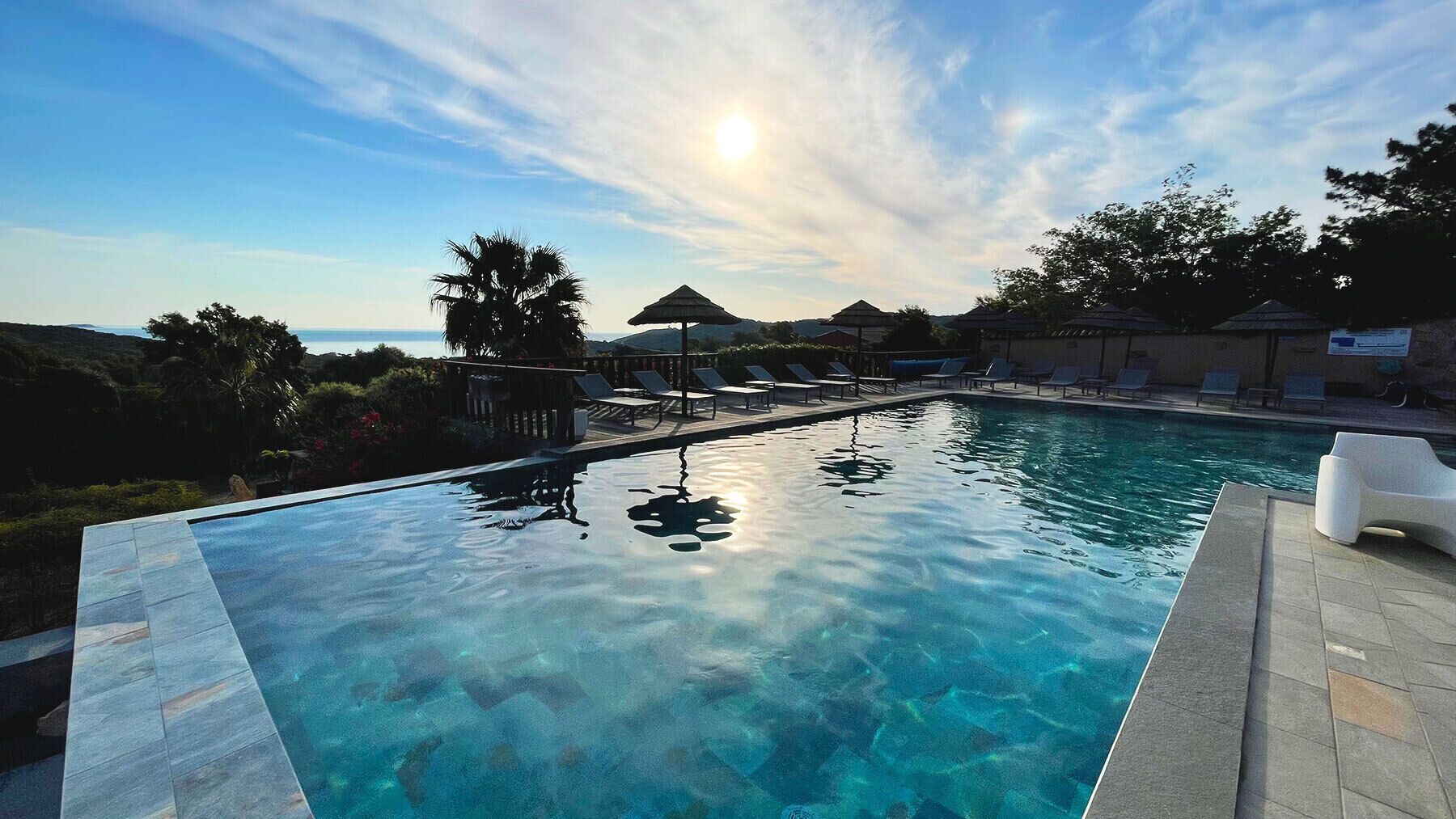 Una piscina azul cristalina con sillas de descanso y sombrillas, rodeada de vegetación y vista al océano.