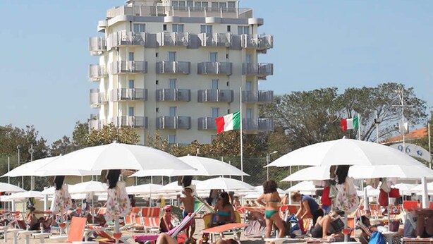 Una playa con tumbonas y sombrillas, un edificio alto con balcones y una bandera.