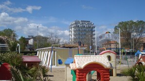 Zona infantil con una casita pequeña roja y beige, rodeada de arena y plantas de palma.