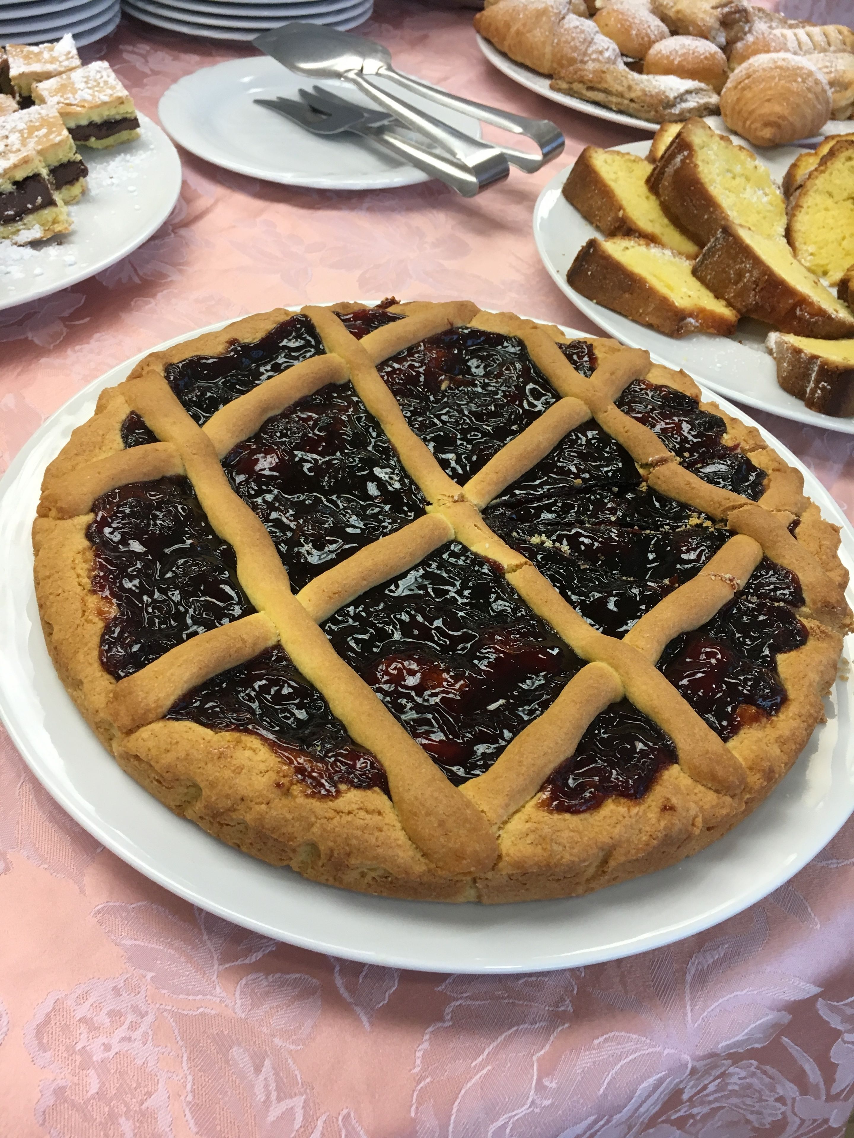 A round pie with a lattice crust on a white plate, surrounded by other desserts and plates on a table with a floral tablecloth.