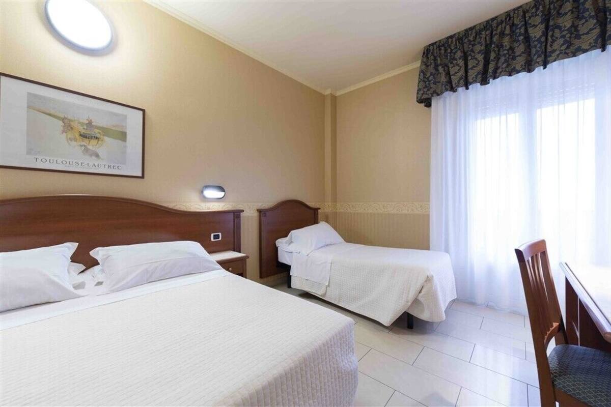 Habitación de hotel con dos camas, cabecera de madera, mesita de noche, una silla y una ventana con cortinas translúcidas.