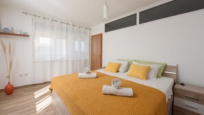 A modern bedroom with a large bed, a wooden nightstand, and a window with sheer curtains. - Sunset Beach Suites (Zadar)