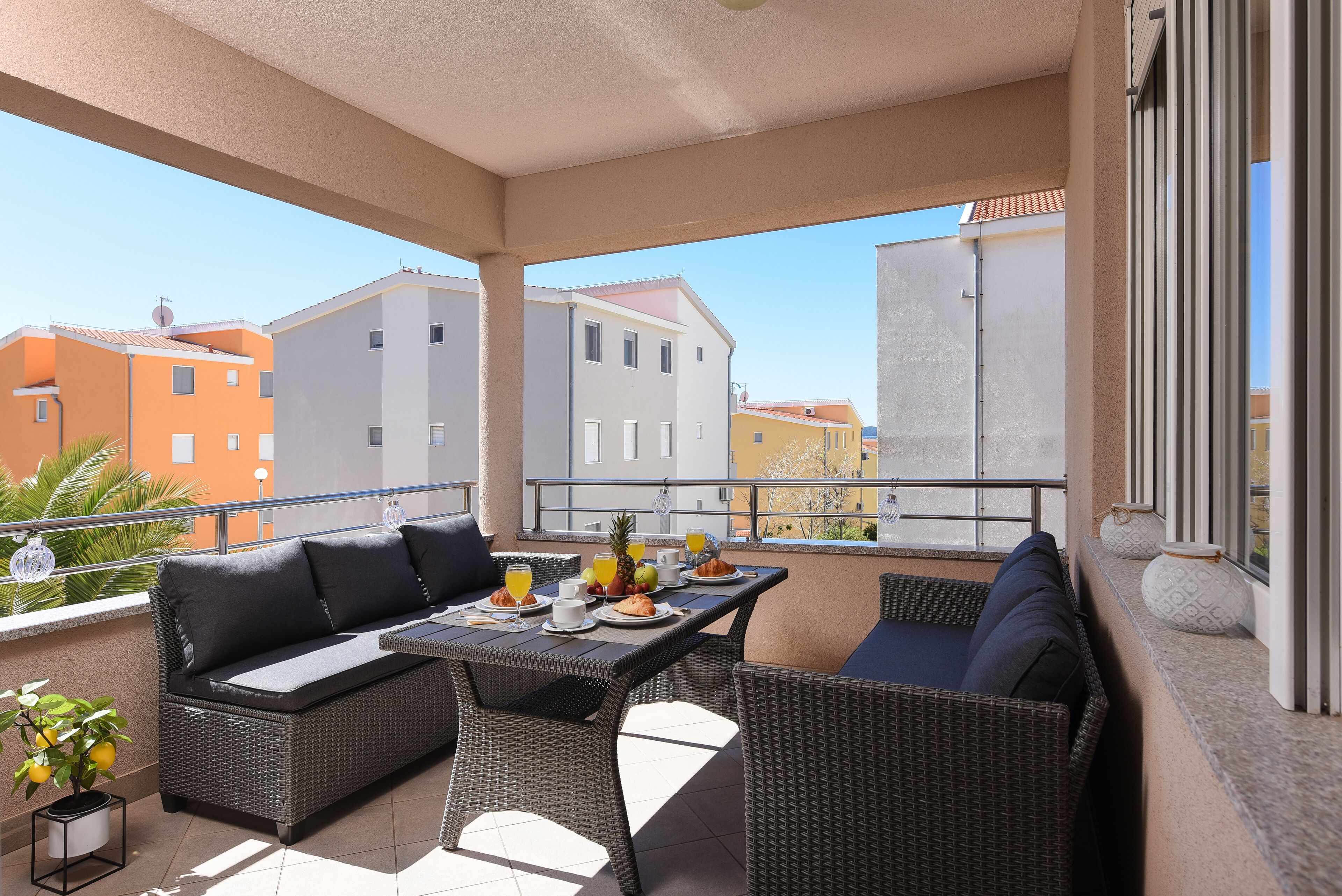 A balcony with wicker furniture, a table with breakfast items, and a view of surrounding buildings.