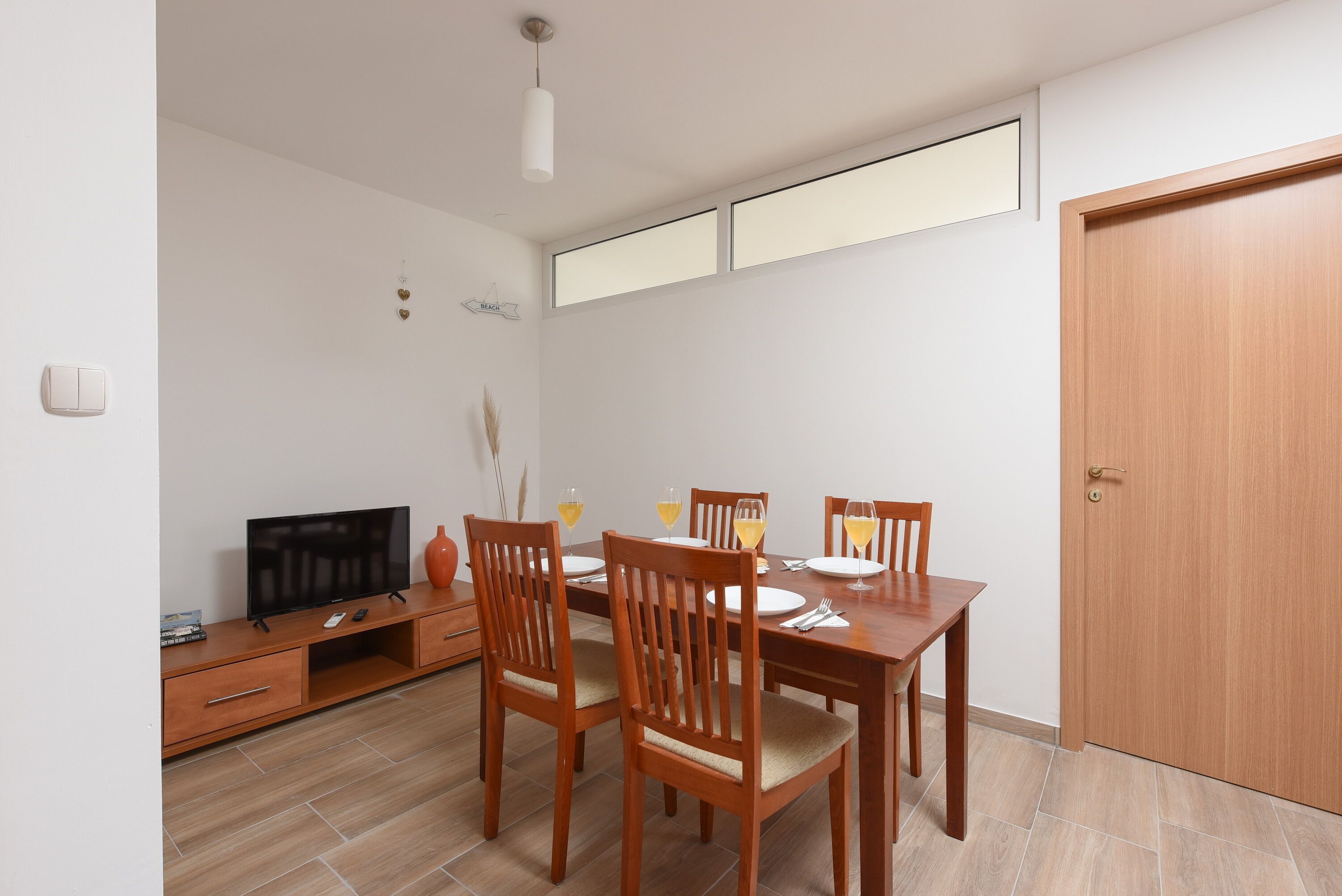 A dining area with a wooden table, four chairs, a television on a wooden cabinet, and a door to the right.