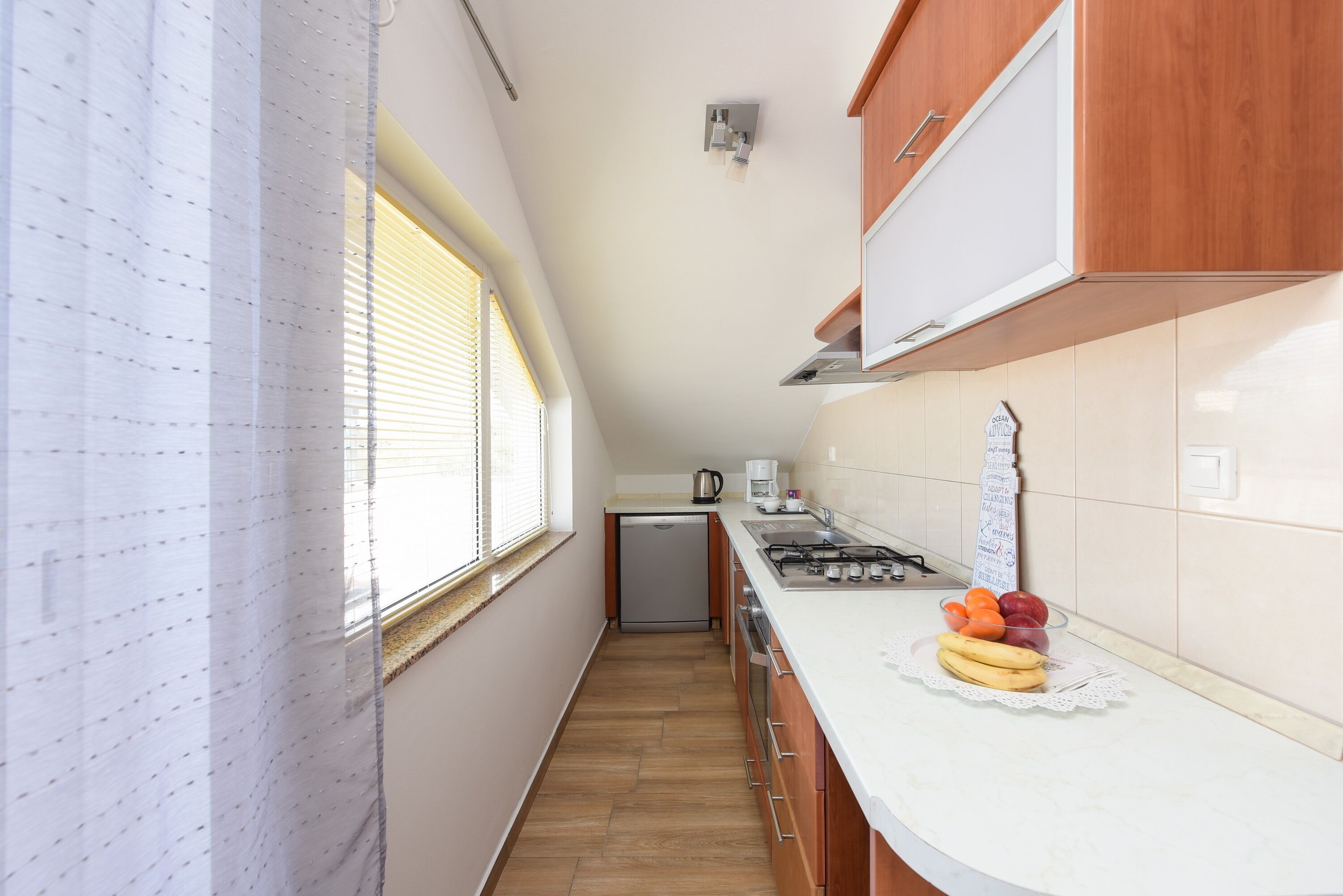 Een smalle keuken met houten kastjes, een wit aanrecht en een raam met jaloezieën.
